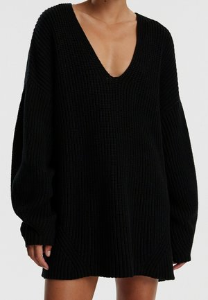Personne portant une robe-pull en maille noire oversize avec un profond décolleté en V et des manches longues, debout devant un fond clair uni.