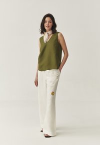 Top fără mâneci de culoare verde măsliniu cu decolteu în V; pantaloni albi cu picioare largi, având broderie cu flori galbene; modelul poartă o eșarfă deschisă.