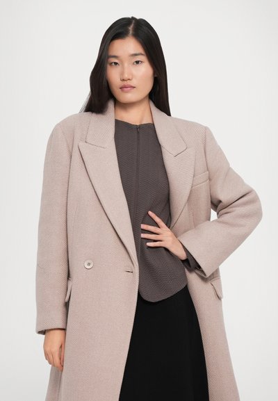 Woman with long straight hair wearing a beige oversized coat over a textured brown top and black skirt, standing with one hand on her waist.