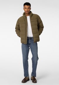 Olivenfarget pufferjakke med høy krage, quiltet design, båret over en hvit t-skjorte, sammen med blå jeans og brune støvler.