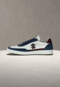 Baskets basses blanches avec des accents bleu marine et rouge, logo noir sur le côté et semelle blanche, exposées sur une surface claire.