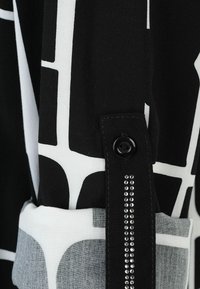 Schwarzes und weißes gemustertes Stoffmaterial mit einer geknöpften Manschette und einer Reihe von dekorativen Strasssteinen entlang des Ärmels. Insgesamt weiche Textur.