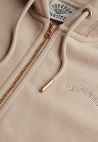 Sweat à capuche beige zippé avec fermeture éclair dorée et cordons de serrage, logo "SUPERDRY" brodé sur la poitrine, et étiquette varsity à l'intérieur du col.