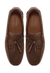 Mocasines de ante marrón con detalles de costura y un lazo atado en la parte delantera. Textura suave y forma redondeada, diseñados para la comodidad.
