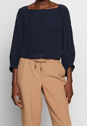 Femme portant un chemisier bleu marine à manches longues avec un col carré et un pantalon beige taille haute avec une ceinture à cordon.