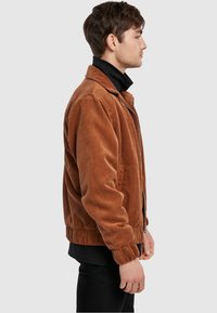 Brown corduroy bomber jacket with a zip front, ribbed cuffs, and collar. Features a textured surface and slightly oversized fit.