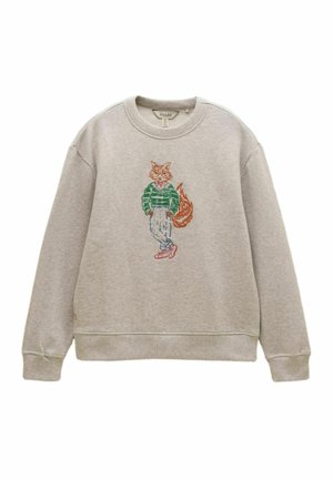 Lysegrå sweatshirt med broderet ræv iført grøn sweater, beige bukser og røde sko, stående med krydsede ben.