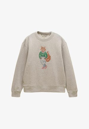 Lysegrå sweatshirt med broderet ræv iført grøn sweater, beige bukser og røde sko, stående med krydsede ben.