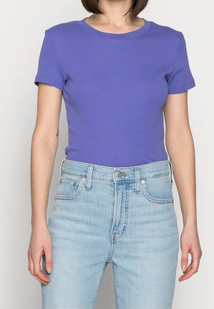 Personne portant un t-shirt violet ajusté à manches courtes rentré dans un jean bleu clair taille haute sur un fond uni.