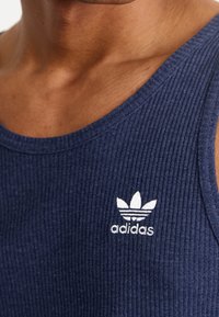 Camiseta sin mangas de canalé azul marino con escote redondo y el logo blanco de Adidas bordado en el lado izquierdo del pecho. Textura de algodón suave.
