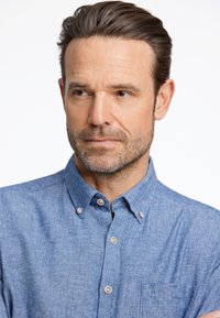 Camicia azzurro chiaro con bottoni, realizzata in un mix di lino, presenta un colletto, una tasca frontale e un tessuto texturizzato con dettagli di cucitura visibili.