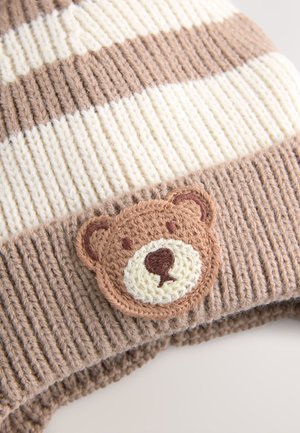 Dryžuota megzta kepurė šviesiai rudos ir kreminės spalvų su tekstūruotu raštu; pasižymi apvaliu meškiuko aplikacija švelniai rudos ir kreminės spalvos siuvinėjimu.