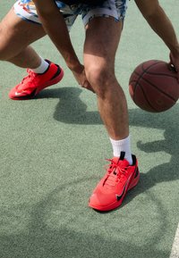 Baskets de sport rouges avec des accents noirs et des lacets blancs, portées avec des chaussettes blanches sur un terrain de basket texturé. Ballon à la main.