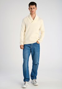 Cremfarbener gerippter Pullover mit Schalkragen, kombiniert mit blauen geraden Jeans und weißen Sneakers. Das Outfit zeichnet sich durch ein lässiges Design aus.