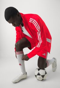 Rød atletikjakke med hvide striber, hvid t-shirt, sokker med røde striber, hvide fodboldstøvler, knælende ved siden af en sort-hvid fodbold.