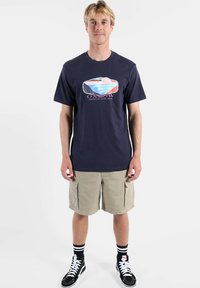 T-shirt en coton marine avec un motif surf multicolore, associé à un short cargo kaki et des baskets noires à lacets blancs.