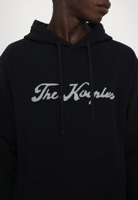 Osoba s tmavou pletí, oblečená v černém mikině s kapucí, na které je bíle vyšitý nápis "The Kooples" přes hrudník, stojící bokem.