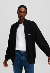 Juodas megztas zip-up džemperis su aukštu apykakle, puošiamas baltu Karl Lagerfeld Jeans logotipu, derinamas su baltu marškinėliu ir mėlynomis džinsomis.