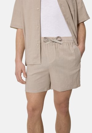 Hombre con pantalones cortos de lino beige con cordón y camisa de manga corta abotonada a juego sobre una camiseta blanca.