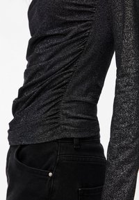 Svart långärmad topp med rynkad design, som har en glittrig yta och figurnära silhuett. Bärs med svarta byxor som har fickdetaljer.