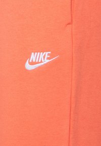 Korálově oranžové tričko Nike s krátkým rukávem, kulatým výstřihem a bílým vyšitým logem na levé části hrudi. Hladká textura.
