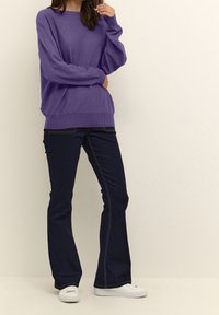 Lila stickad tröja med långa ballongsärmar och ribbat avslut, tillsammans med mörkblå utsvängda jeans med ljusa sömmar och vita sneakers.