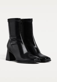 Schwarze Knöchelstiefel aus glänzendem Leder, mit einem Reißverschluss an der Vorderseite, einer quadratischen Zehenform und einem massiven Absatz. Glatte Textur mit minimalen Nähdetails.