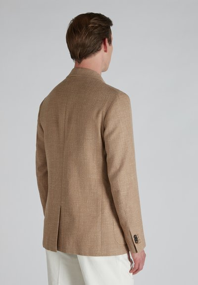 Blazer texturizado en tono beige con corte entallado, que cuenta con una abertura trasera sencilla y detalles de botones en las mangas. Fondo liso.