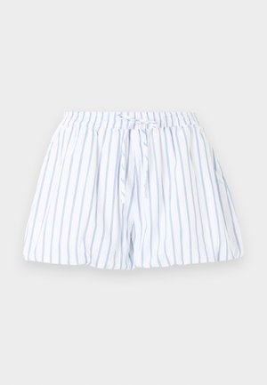 Femme portant une chemise blanche boutonnée et un short assorti à rayures bleues, debout les jambes croisées, portant des ballerines noires à pois blancs.