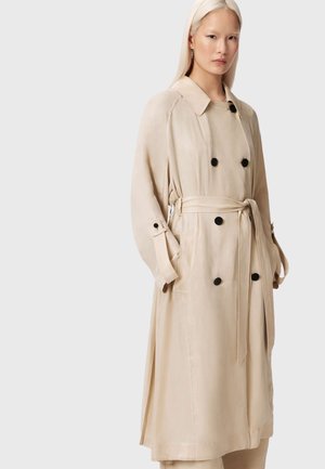 Kvinde med langt blondt hår iført en beige dobbeltradet trenchcoat med sorte knapper og bælte, stående foran en ensfarvet lys baggrund.