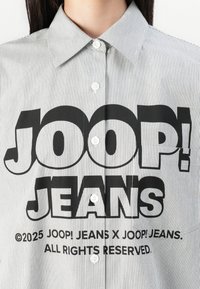 Stripete skjorte med knapp i svart og hvitt, med et stort grafisk trykk av "JOOP! JEANS" på forsiden. Inkluderer hvite knapper.