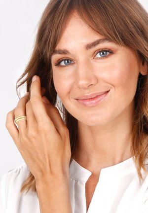 Gold-tone ring worn on the right hand, features a double band design. The subject has light brown hair and wears a white blouse with a collar.