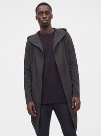 Young man wearing a dark long hooded cardigan over a black t-shirt and black pants, standing against a plain light background.