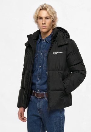 Ung mand iført en sort Karl Lagerfeld Jeans dunjakke over en blå denimskjorte og jeans med brunt bælte, stående mod hvid baggrund.