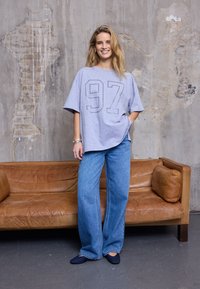 Hall suurendatud t-särk, millel on ees print number 97, kombineeritud laia säärega siniste teksade ja tumedate libisevate jalanõudega.