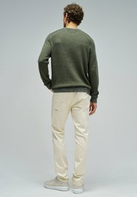 Zielony sweter z dzianiny z prążkowanymi mankietami, w połączeniu z kremowymi spodniami o slim fit. Szare sneakersy dopełniają stylizację, widoczna z tyłu.