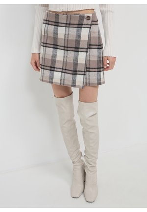 Frau trägt einen beige-weiß karierte Minirock mit Knopfdetail, kombiniert mit einem weißen Rippsweater und weißen kniehohen Stiefeln.