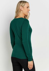 Grünes Langarmshirt mit runden Ausschnitt und tailliertem Schnitt. Der Stoff ist glatt und die Ärmel reichen bis zu den Handgelenken.
