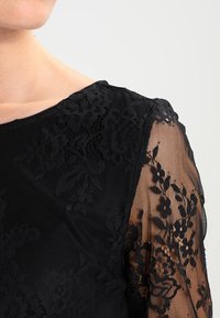 Abito nero con sovrapposizione di pizzo floreale, maniche lunghe trasparenti, scollo rotondo, tessuto strutturato, dettagli intricati sulle maniche.