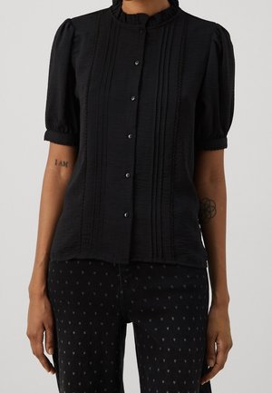 Mujer vestida con una blusa negra de manga corta con cuello con volantes y detalles de pliegues, combinada con pantalones negros que presentan pequeños patrones de estrellas.