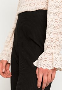 Top en dentelle blanche avec un motif floral et des manches à volants, associé à un pantalon noir côtelé taille haute. Textures douces et couleurs contrastées.