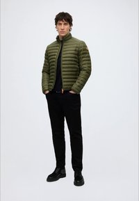 Homme debout, mains dans les poches, portant une veste matelassée vert olive, pantalon noir, chemise noire et chaussures noires sur un fond uni.