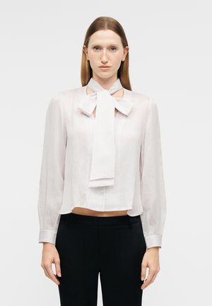 Kobieta o prostych brązowych włosach, ubrana w białą bluzkę z długim rękawem z dużą kokardą pod szyją i czarne spodnie, stojąca na jednolitym tle.