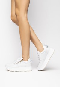 Hvide lædersneakers med en tyk hvid sål, der har en sidelukning med lynlås, snørebåndsdesign og en lille sløjfedetalje på hælen.