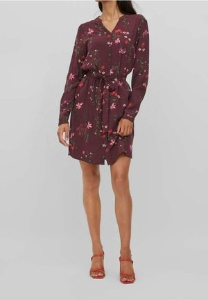 Kobieta w długiej do kolan sukience z długim rękawem w kolorze fioletowym z różowym kwiatowym wzorem i wiązaniem w talii, w parze z czerwonymi sandałami na wysokim obcasie z paskami.