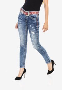 Schlanke Denim-Jeans in Hellblau mit verwaschenen Details. Mit silbernem Nähen und einem mit Juwelen besetzten roten Gürtel. Getragen mit schwarzen High Heels.