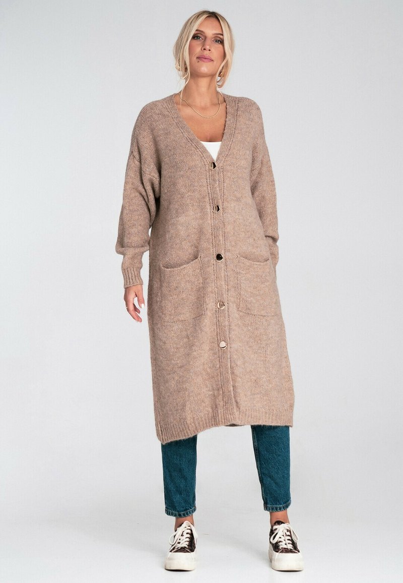 Kobieta ubrana w długi beżowy kardigan zapinany na guziki, nosząca niebieskie dżinsy i trampki w cętki, stojąca na tle jasnego, jednolitego tła.