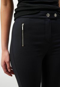 Pantaloni negri ajustați, fabricați dintr-un material neted, care prezintă un buzunar cu fermoar pe lateral, două butoane metalice la talie și o croială adaptată.