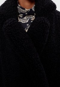 Abrigo negro y peludo con una textura suave y un cuello ancho, superpuesto a una camisa oscura con un patrón que presenta motivos de dragones en colores claros.