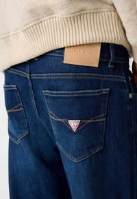 Tumed sinised denim teksad tekstuursete pindadega. Omavad tagakotisid kolmnurkse GUESS logoga ja pruunist nahast sildist vöökohal.